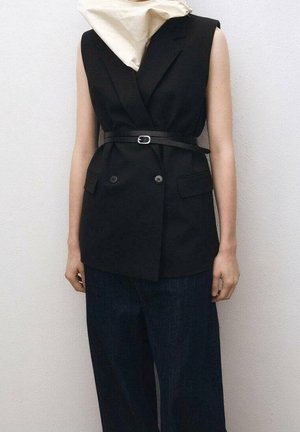 Czarna bezrękawnikowa marynarka z podwójnymi guzikami i paskiem, noszona do luźnych ciemnych spodni, zestawiona z kremowym szalikiem na szyi.