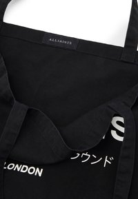 Black fabric tote bag with wide straps, featuring white text designs and a branded label on the top edge. Smooth texture, minimalistic design.
