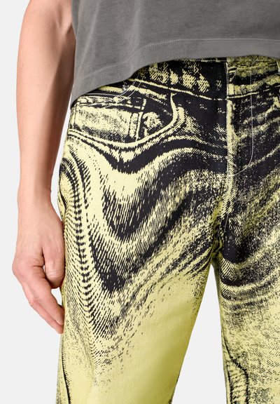 High-waisted denim jeans featuring a bold black and yellow abstract pattern, with a slim fit and five-pocket design, shown at the waist.