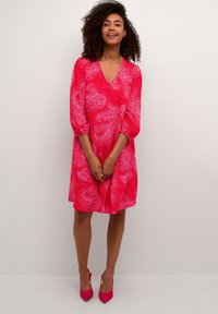 Röd klänning med paisley-mönster, trekvartslånga ärmar och V-ringning, tillverkad av lätt material. Matchas med rosa högklackade skor.