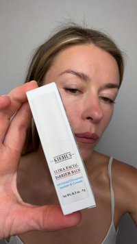 Woman holding a white box of Kiehl's Ultra Facial Barrier Balm close to her face against a plain background.