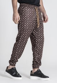 Pantalones de chándal marrones con un diseño estampado y cintura elástica, que presentan un diseño de logotipo repetido en color beige. Combinados con zapatillas negras con suela blanca.