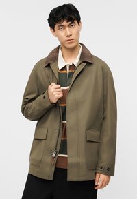 Junger Mann trägt eine olivgrüne Jacke mit braunem Cordkragen über einem gestreiften Hemd und hält den Reißverschlussbereich der Jacke.