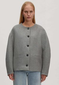 Frau trägt eine graue Wolljacke mit Knöpfen und zwei vorderen Taschen sowie eine hellblaue Jeans, steht vor einem einfarbigen Hintergrund.