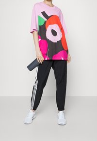 Mujer con camisa rosa floral de gran tamaño, pantalones deportivos negros con rayas blancas, zapatillas blancas, sosteniendo una esterilla de yoga cilíndrica y oscura.