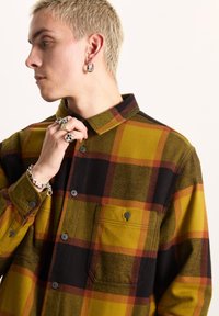 Camicia in flanella con un motivo a quadri giallo, nero e arancione, design con bottoni, taschino sul petto e bottoni neri. Tessuto texture evidente.