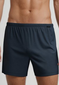 Mannelijk bovenlijf in eenvoudige donkerblauwe elastische shorts, staand tegen een neutrale lichte achtergrond.