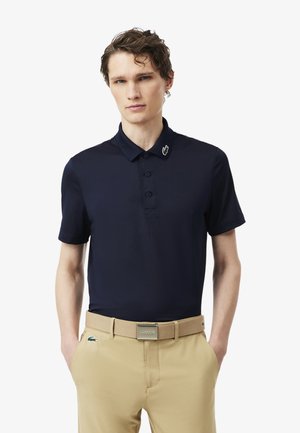 Joven con camiseta polo azul marino y un pin de cocodrilo verde en el cuello, pantalones beige y cinturón beige con hebilla metálica.