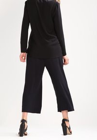 Top nero a maniche lunghe abbinato a pantaloni neri a gamba larga. Sandali con tacco alto presentano cinturini alla caviglia. Tessuto liscio con design minimale.
