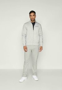 Grå lynlås sweatshirt og joggers i et blødt stof, med et logo på brystet og buksebenet, parret med hvide sneakers.
