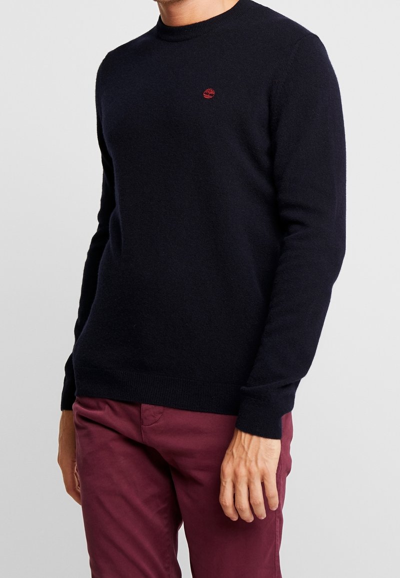 Homme portant un pull noir ajusté à col rond avec un petit logo rouge sur la poitrine et un pantalon bordeaux, se tenant devant un fond uni.