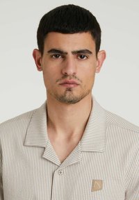 Camisa de botones a rayas beige con una textura suave, que presenta un cuello y un pequeño parche con el logo en el lado izquierdo.