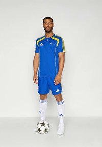 Camiseta de fútbol azul con detalles en amarillo, pantalones cortos azules a juego y calcetines blancos. Logotipo de Adidas mostrado de forma prominente. Balón de fútbol blanco y negro.
