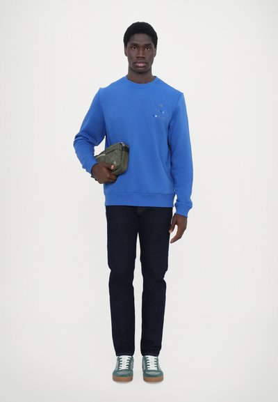Sweatshirt bleu à col rond avec un logo subtil, jeans foncés et baskets vertes avec des lacets blancs ; tenant un sac à pochette olive.