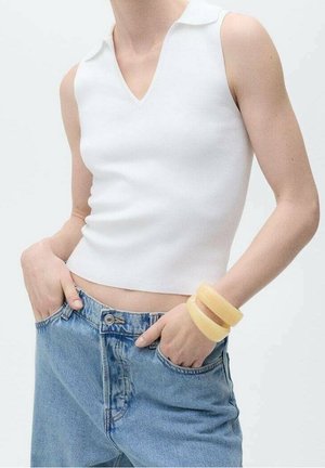 Personne portant un haut blanc sans manches à col en V, un jean en denim bleu, et deux bracelets jaunes au poignet gauche, les mains dans les poches.