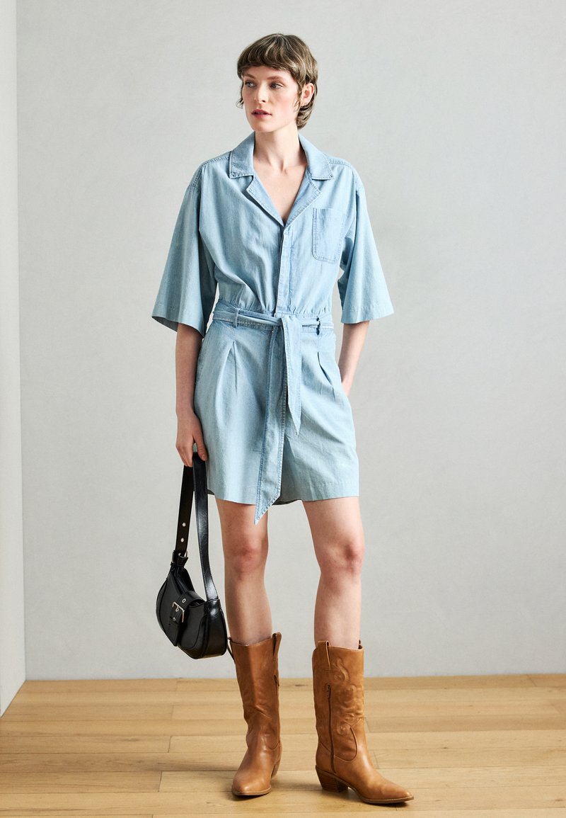 Φόρεμα από τζιν ανοιχτό μπλε με κοντά μανίκια, ζώνη στη μέση και μπροστινή τσέπη. Συνδυασμένο με ψηλές καφέ καουμπόικες μπότες και μαύρη τσάντα ώμου.