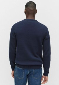 En man i en marinblå stickad tröja och blå jeans står vänd bort från kameran mot en enkel ljusgrå bakgrund.