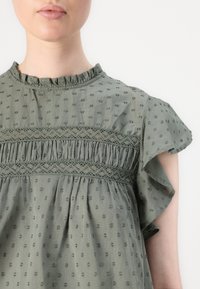 Lys grøn top med et tekstureret prikkemønster, flæser på ærmerne og en smocket blonde detalje over brystet. Blødt bomuldsstof.