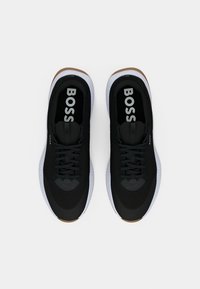 Un paio di scarpe da ginnastica nere con suole intermedie bianche, suole esterne marroni e il marchio "BOSS" all'interno dell'area del tallone, viste dall'alto.