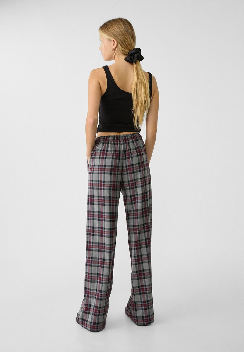 Kobieta z długimi blond włosami związanymi czarną gumką, ubrana w czarny top na ramiączkach i szare szerokie spodnie w kratę, stoi z rękami w kieszeniach.