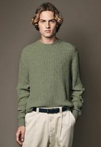 Pull à côtes vert avec des manches longues et un col rond ; associé à un pantalon beige et une ceinture bleu marine avec des coutures blanches.