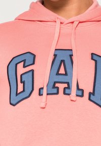 Sudadera de color rosa claro con un bolsillo frontal, capucha con cordones y bordado azul con la letra "G.A.P.". Tejido suave con una textura lisa.