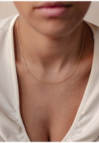 Collier pendentif en or sur une peau nue, avec une chaîne fine et lisse. L'arrière-plan présente un tissu clair texturé avec des plis subtils.
