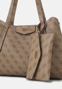 Sac fourre-tout en toile marron avec garnitures dorées, arborant le logo Guess, un motif décoratif, et un porte-monnaie assorti attaché par un fermoir.