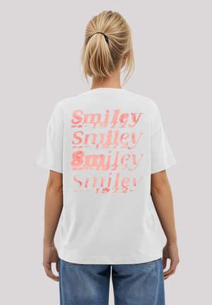 White cotton t-shirt featuring a repeating pink text design reading "Smiley EST 1972" across the back, relaxed fit, short sleeves.