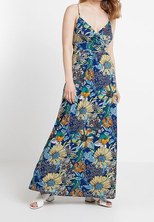 Kobieta w niebieskiej sukience maxi w kwiaty z cienkimi ramiączkami i białych sandałach na słupku z odkrytymi palcami, stojąca na jednolitym tle.