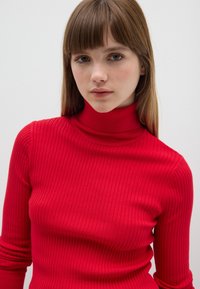 Rode geribde coltrui met lange mouwen, voorzien van een aansluitende pasvorm en verticale textuur. Effen achtergrond.