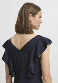 Marindress med V-ryggdesign, ruffled ärmar och en texturerad tyg. Baksidan avslöjar sammetslen hud längs halslinjen.