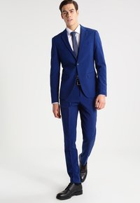 Blå skräddarsydd kostym med slim fit, kavaj med en knapp, byxor med slät front och vit skjorta. Komplett med en mönstrad slips och svarta skor.