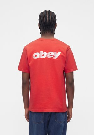 Person med flettet hår står med fronten væk, iført en rød T-shirt med hvid "obey" tekst på bagsiden og blå jeans.