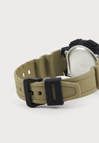 Bracelet de montre en silicone kaki avec un design texturé. Équipé d'une boucle en métal noir et d'un boîtier ovale en argent avec une lunette noire.