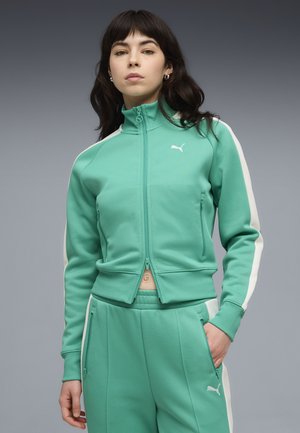 Jonge vrouw in een turquoise Puma-trainingspak met witte strepen, staand tegen een effen grijze achtergrond met een neutrale uitdrukking.