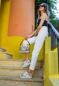 Weiße Sneakers mit schwarzen und roten grafischen Designs, kombiniert mit weißen Hosen und einer gemusterten Handtasche, vor einem bunten Hintergrund.