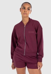 Sweatshirt bordeaux zippé avec bords côtelés et détail de logo, associé à un short coordonné. Design doux et décontracté, idéal pour une tenue de loisirs.