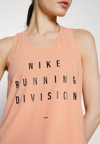 Top in pesca con testo nero "NIKE RUNNING DIVISION". Presenta un collo rotondo e un design senza maniche. Tessuto dalla texture liscia.
