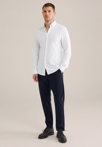 Balta garām piedurknēm krekls ar pogājamu apkakli, kombinēts ar tumši ziliem biksēm un melniem ādas apaviem, uz neitrāla fona.