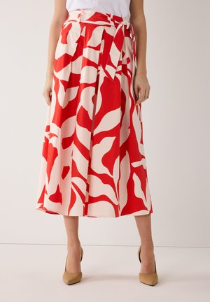 Une femme portant une jupe mi-longue rouge et blanche à motifs, avec une ceinture nouée, et des talons pointus beiges.