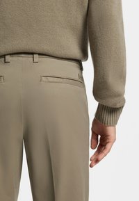 Beige Hosen mit glatter Textur und Seitentaschen, kombiniert mit einem hellbraunen, gerippten Wollpullover. Dezente Steppnähte an der Hose.