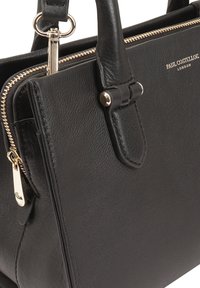 Sac à main en cuir noir avec fermeture éclair dorée, couture saillante, poignées supérieures et une étiquette détachable. Présente une finition texturée et un design minimaliste.