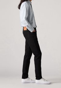 Person med svarta slim jeans, vita sneakers och en blå och vit randig skjorta med knappar som står på sidan med händerna i fickorna.