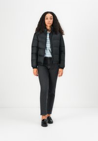 Sort pufferjakke med quiltet design og høj krave, parret med grå jeans og sorte loafers, båret over en lysstribet skjorte.