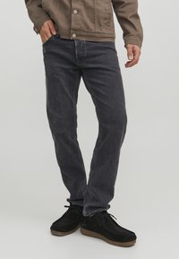 Jeans in denim grigio scuro con vestibilità dritta. Tessuto in cotone con una sottile texture, abbinati a scarpe in suede nere e una giacca marrone.
