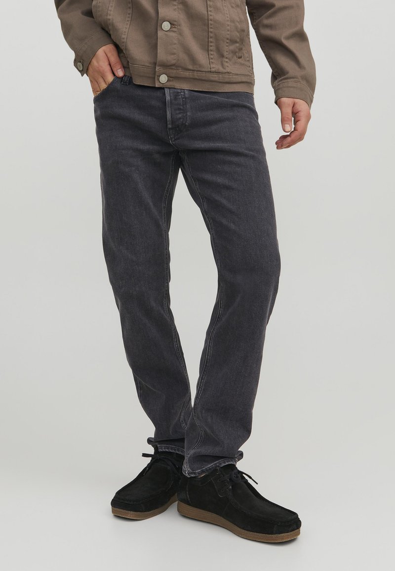 Jeans in denim grigio scuro con vestibilità dritta. Tessuto in cotone con una sottile texture, abbinati a scarpe in suede nere e una giacca marrone.