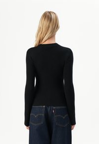 Mulher com longos cabelos loiros, vestindo um suéter preto justo com textura de costela e jeans azul-escuro, mostrada de costas contra um fundo simples.