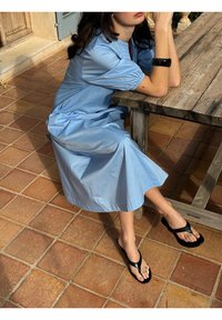 Žena u svijetloplavoj haljini sjedi za drvenim stolom na terakotin pločicama, nosi crne sandale i crnu narukvicu.