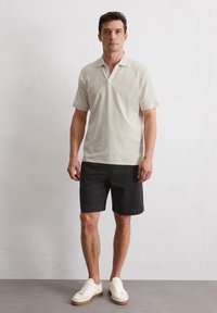 Korte mouwen, lichtbeige polo van zachte stof, gecombineerd met donkere shorts. Witte sneakers met een gestructureerde zool maken de outfit af.